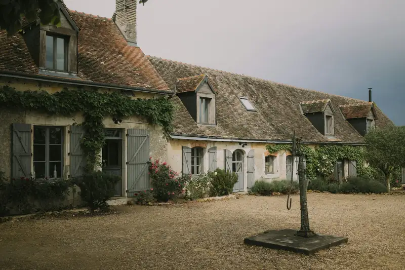 Maison ancienne en pierre aux volets clairs, cour gravillonnée et arbre isolé